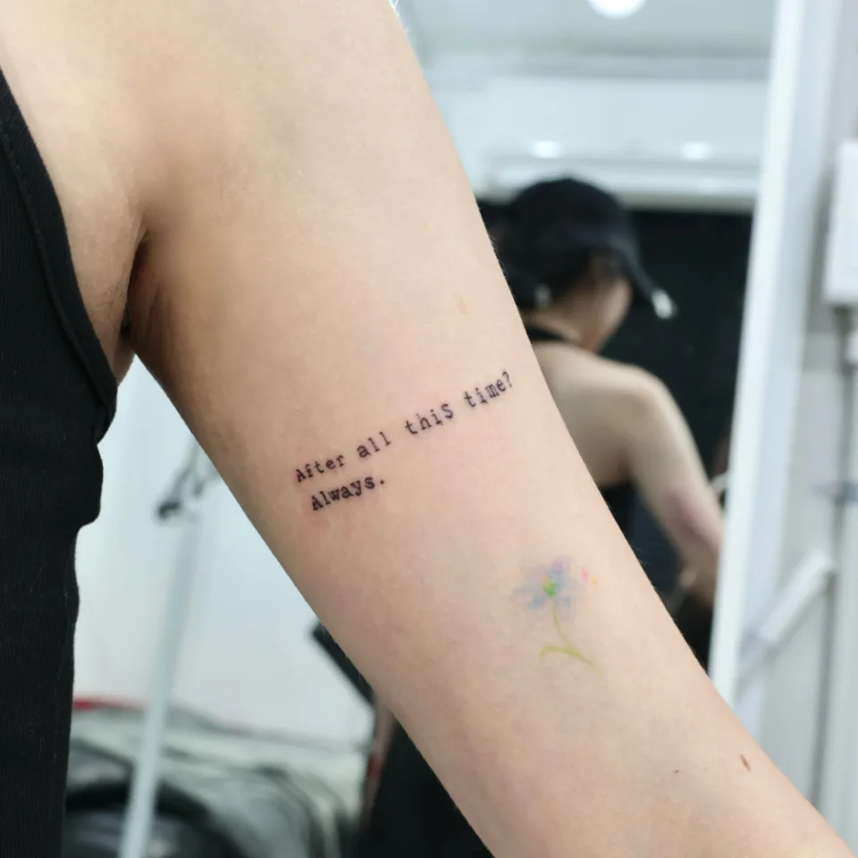같은 위팔 레터링 타투를 멀리서 본 사진, 문장 아래쪽에 별도의 작은 파스텔 꽃 타투가 보임