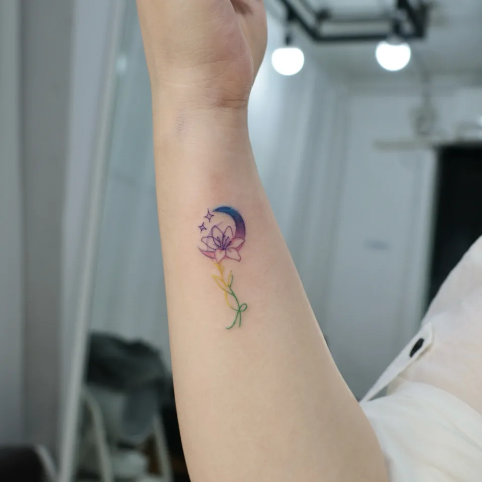 팔을 들어 올려 찍은 같은 손목 타투 사진, 달 위로 꽃이 피고 별 두 개와 노랑·초록 줄기가 보이며 손목 주름과 흰 소매가 함께 보임