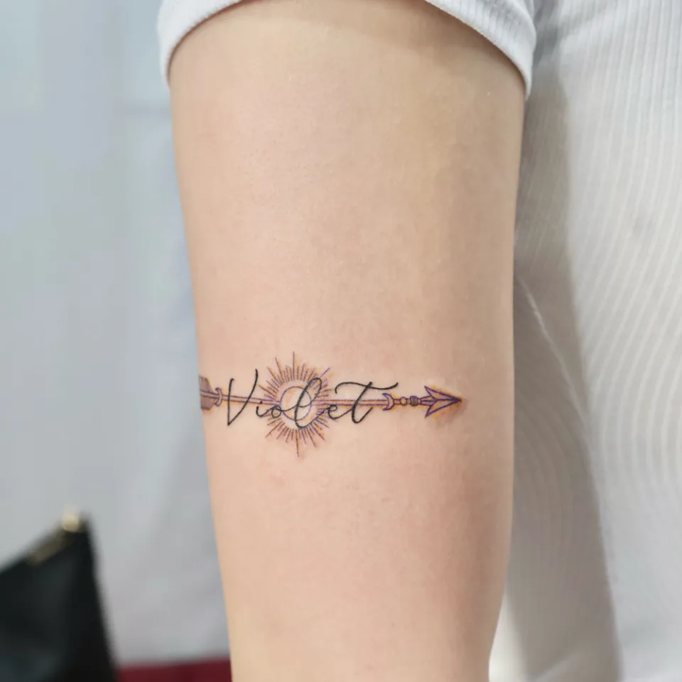 같은 이름 화살 밴드 타투를 위팔 옆에서 본 사진, 화살 깃과 촉이 팔 둘레를 따라 양쪽으로 뻗고 가운데 햇살 원이 보임