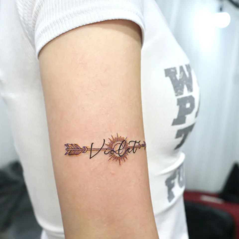 흰 반팔 소매 아래 위팔에 필기체 이름과 화살, 햇살 원이 가로 띠처럼 놓인 타투를 옆모습에서 본 사진