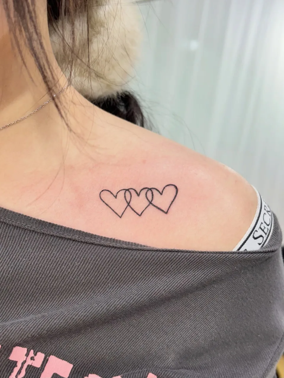 같은 주제의 다른 작업, 쇄골과 어깨 사이에 가는 검은 선으로 그린 하트 세 개가 서로 겹쳐 이어진 타투 사진