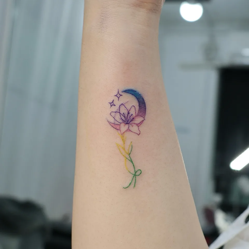 손목 안쪽에 초승달과 꽃, 작은 별 두 개가 있는 컬러 타투 사진, 달은 파랑에서 보라와 분홍으로 번지는 그라데이션이고 줄기는 노랑에서 초록으로 이어지며 끝에 작은 매듭이 있음